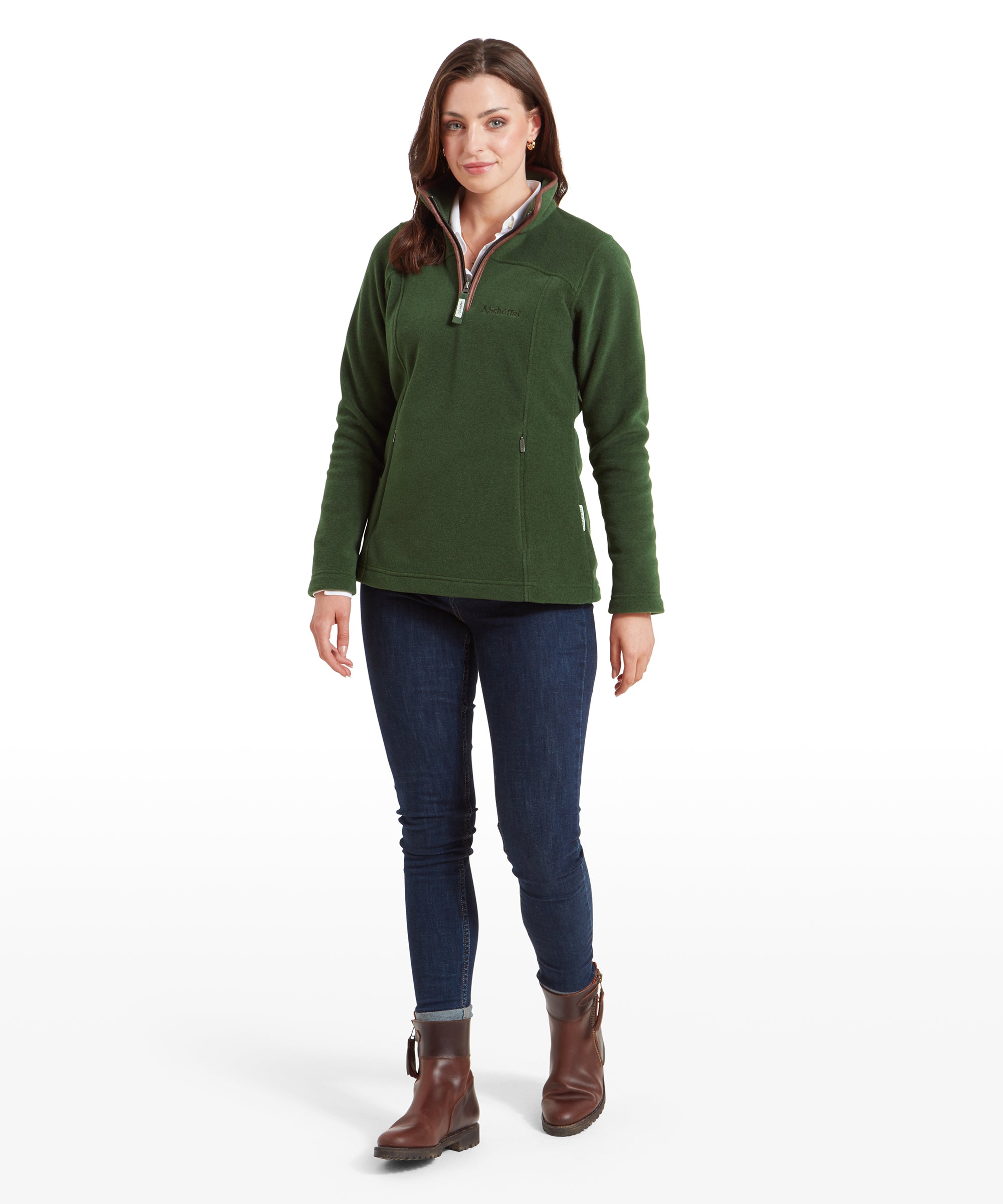 A woman is depicted wearing a Schöffel Tilton 1/4 Zip Fleece for Women in Light Green with brown trim around the collar. The fleece is paired with dark blue skinny jeans and brown ankle boots. The woman has long, dark hair and wears a light blue button-up shirt underneath the fleece. She stands confidently with a slight smile, her left hand at her side and her right hand partially out of the frame.