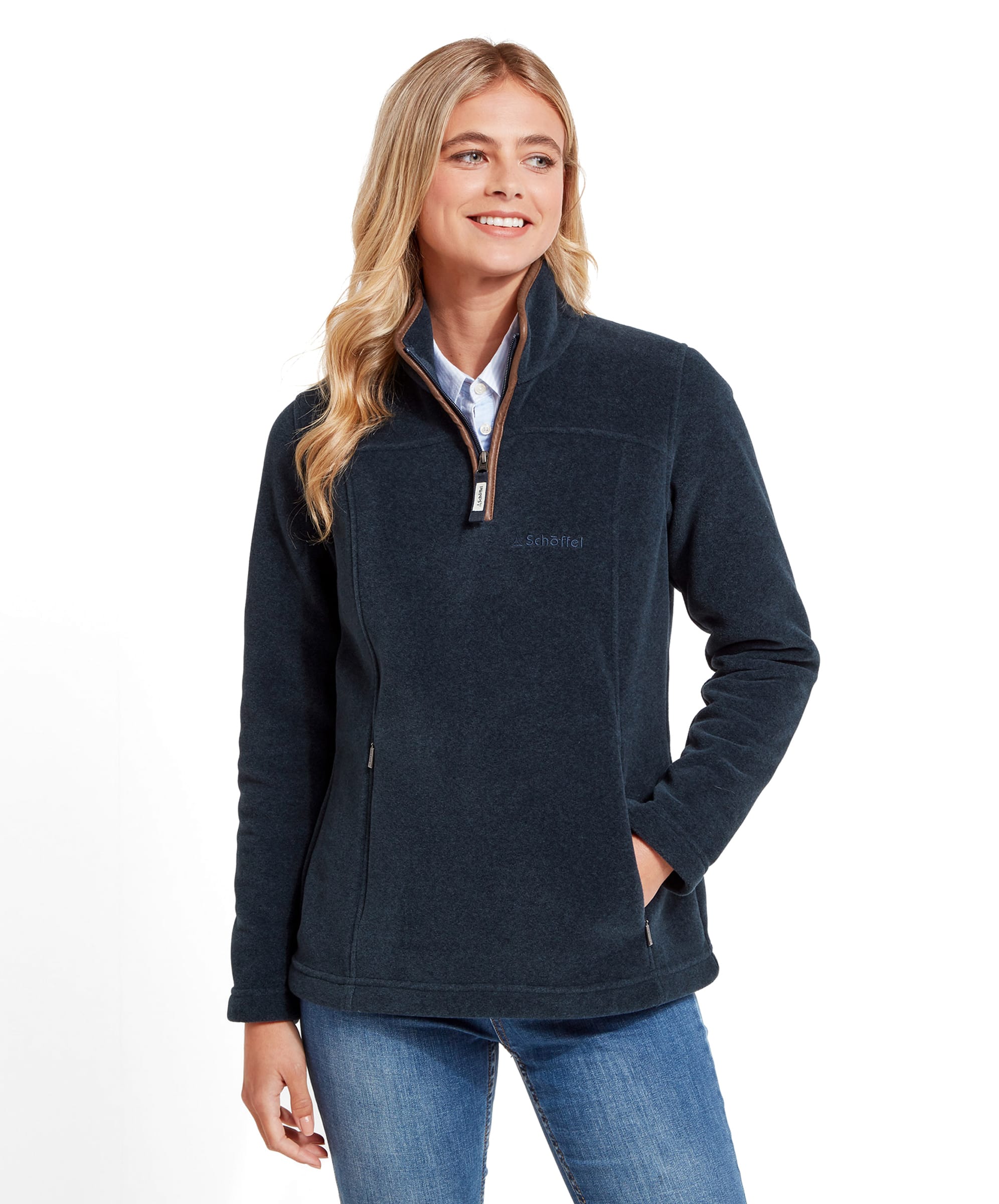 A woman is shown wearing a Schöffel Tilton 1/4 Zip Fleece for Women in Blue with a high collar. The fleece has a small, subtle logo on the left chest area. She pairs the fleece with a light blue button-up shirt underneath, and her blonde hair is styled in loose waves. The woman is smiling softly, with her left hand in her pocket, creating a casual and comfortable look.