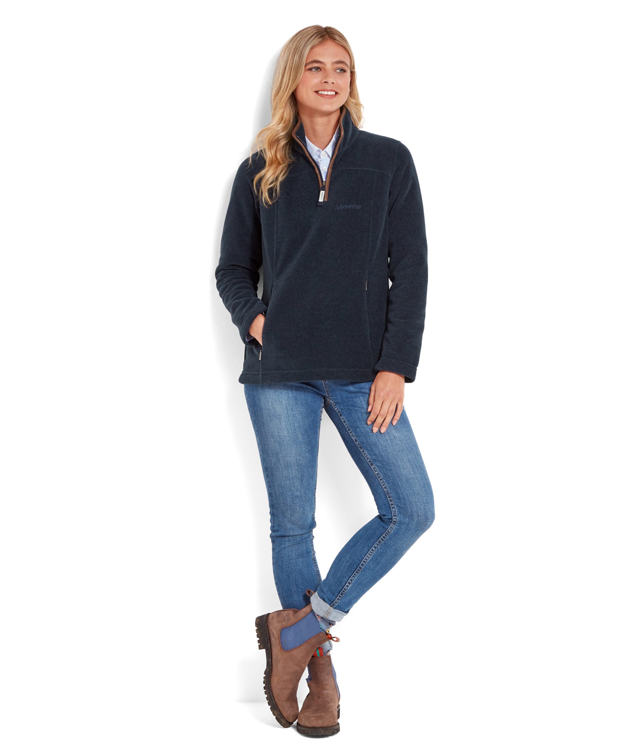 The same woman is depicted in a full-body shot wearing the Schöffel Tilton 1/4 Zip Fleece for Women in Blue, light blue skinny jeans, and brown ankle boots with blue side panels. She stands with one leg slightly crossed over the other, her left hand in her pocket, and smiles slightly while looking off to the side. The outfit presents a relaxed, outdoorsy style.