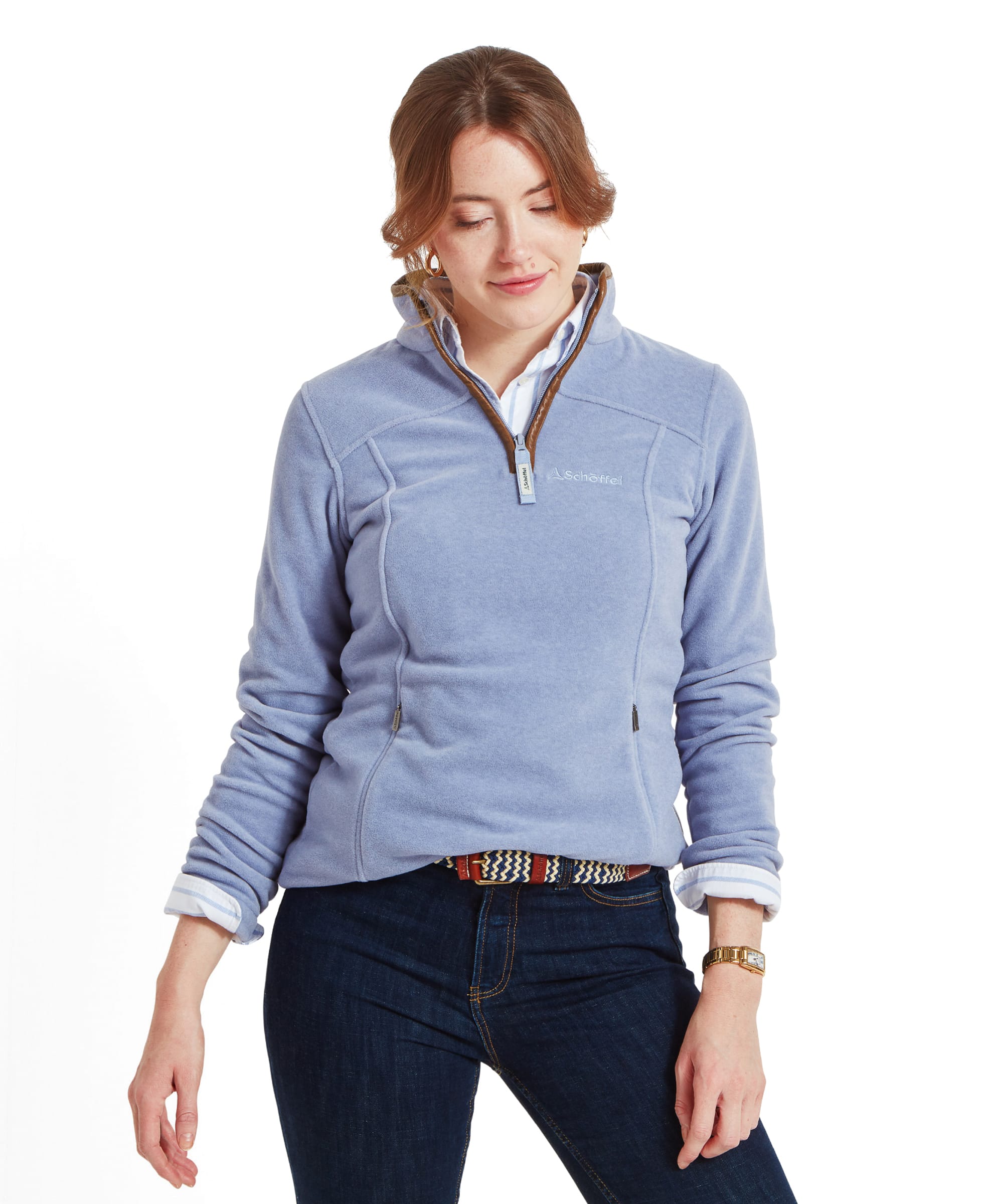 A woman is seen wearing a Schöffel Tilton 1/4 Zip Fleece for Women in Light Blue with brown trim around the collar and zipper. She has her hair styled in a loose, low updo, and wears a striped blue and white button-up shirt underneath. The woman looks downwards with a soft smile, creating a relaxed and casual appearance. She pairs the fleece with dark blue jeans and a woven belt.