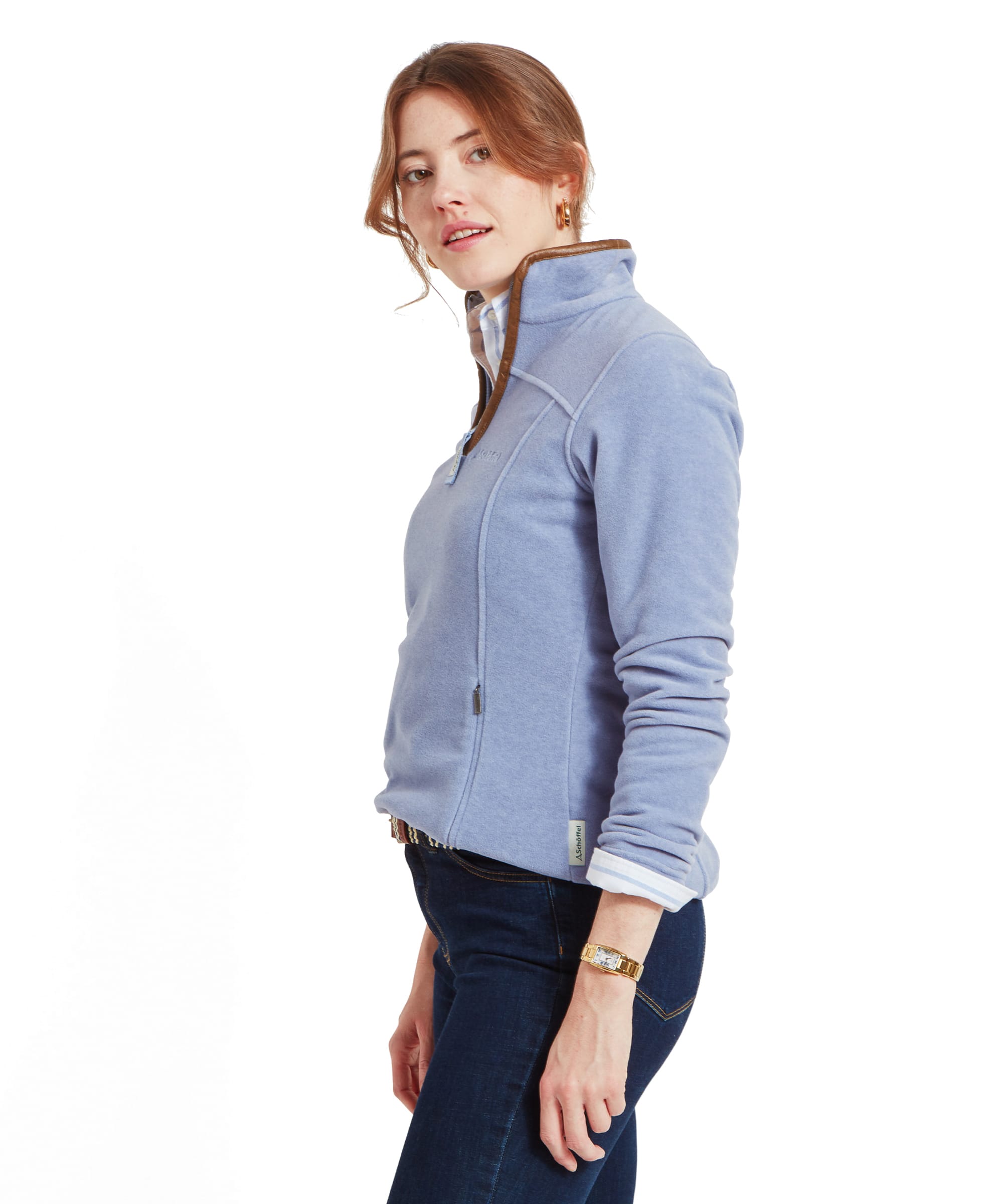 The woman is captured from the side, showing the fit of the Schöffel Tilton 1/4 Zip Fleece for Women in Light Blue. The fleece has a high collar with brown trim, and the fabric appears soft and cozy. She wears dark blue jeans, and a gold wristwatch is visible on her left wrist. Her expression is neutral as she looks slightly over her shoulder.