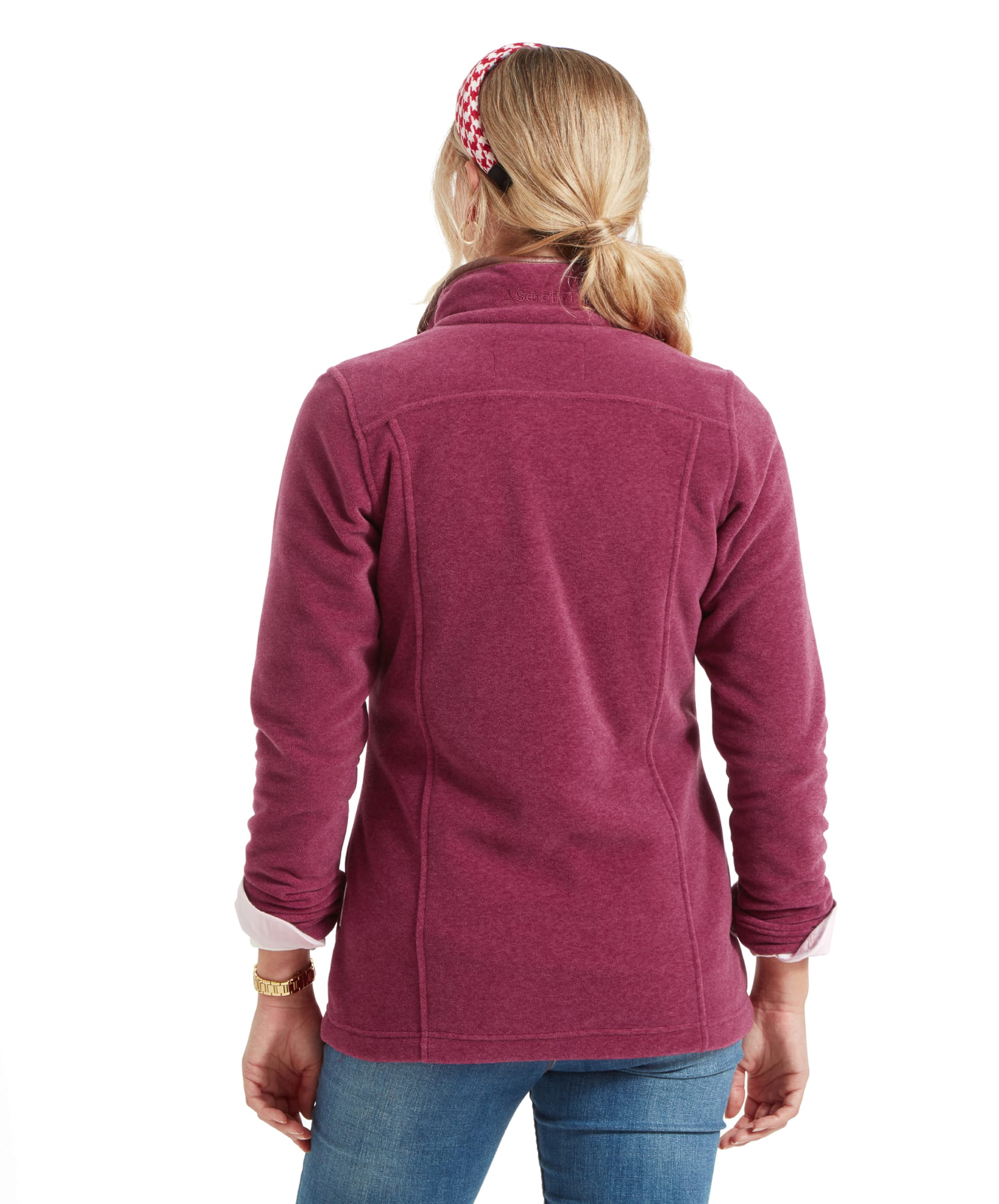 The woman is shown from the back, highlighting the simple and fitted design of the Schöffel Tilton 1/4 Zip Fleece for Women in Pink. The back has subtle seam details, and the collar stands upright. Her blonde hair is tied back in a low ponytail, secured with the red and white headband, which adds a pop of colour to the overall look.