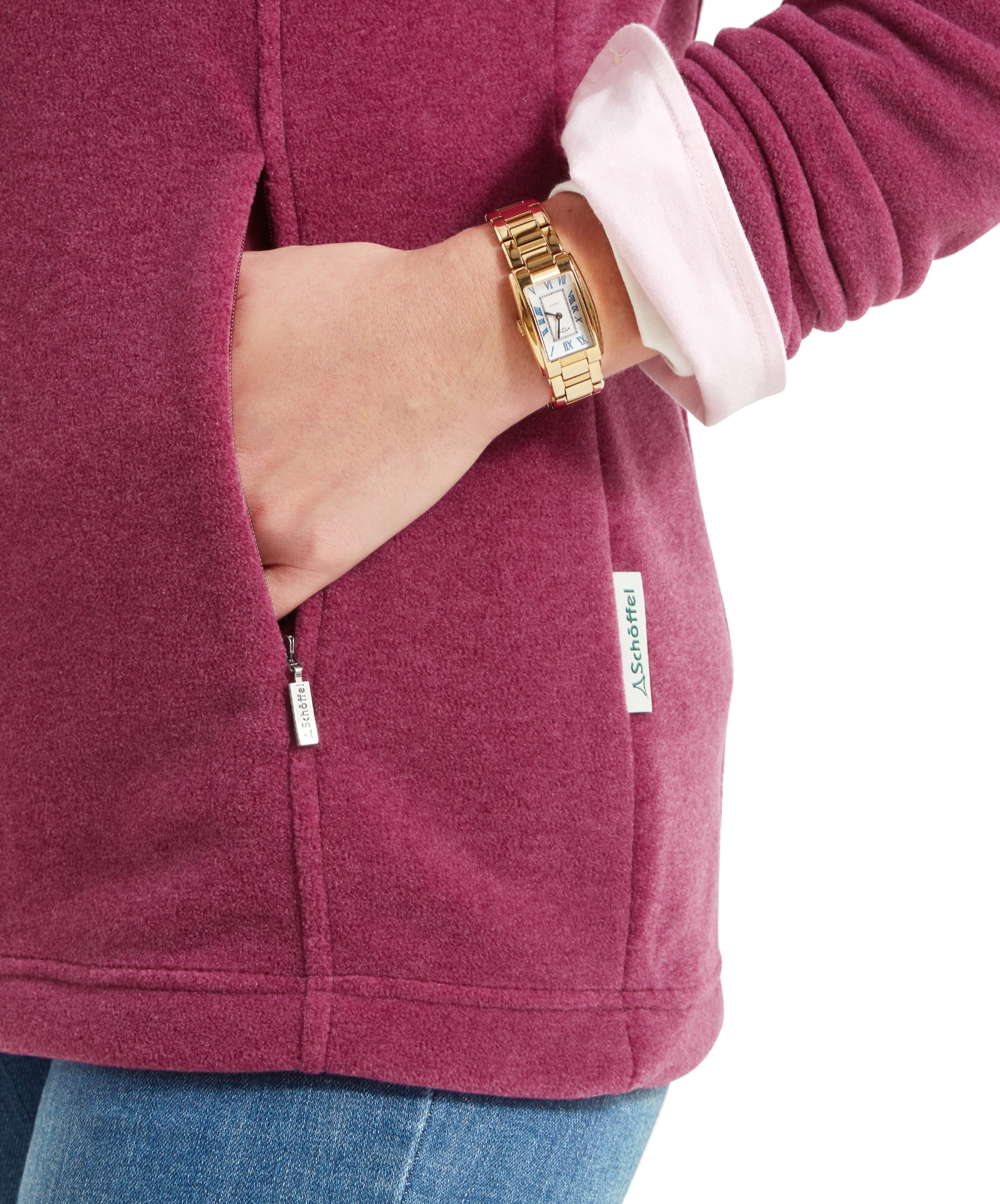 A close-up shot shows the woman’s hand tucked into one of the Schöffel Tilton 1/4 Zip Fleece for Women in Pink’s side pockets. The deep pink fabric contrasts with her light blue jeans, and the brand’s logo tag is visible near the pocket. The woman is wearing a gold wristwatch, adding a touch of elegance to the casual outfit.