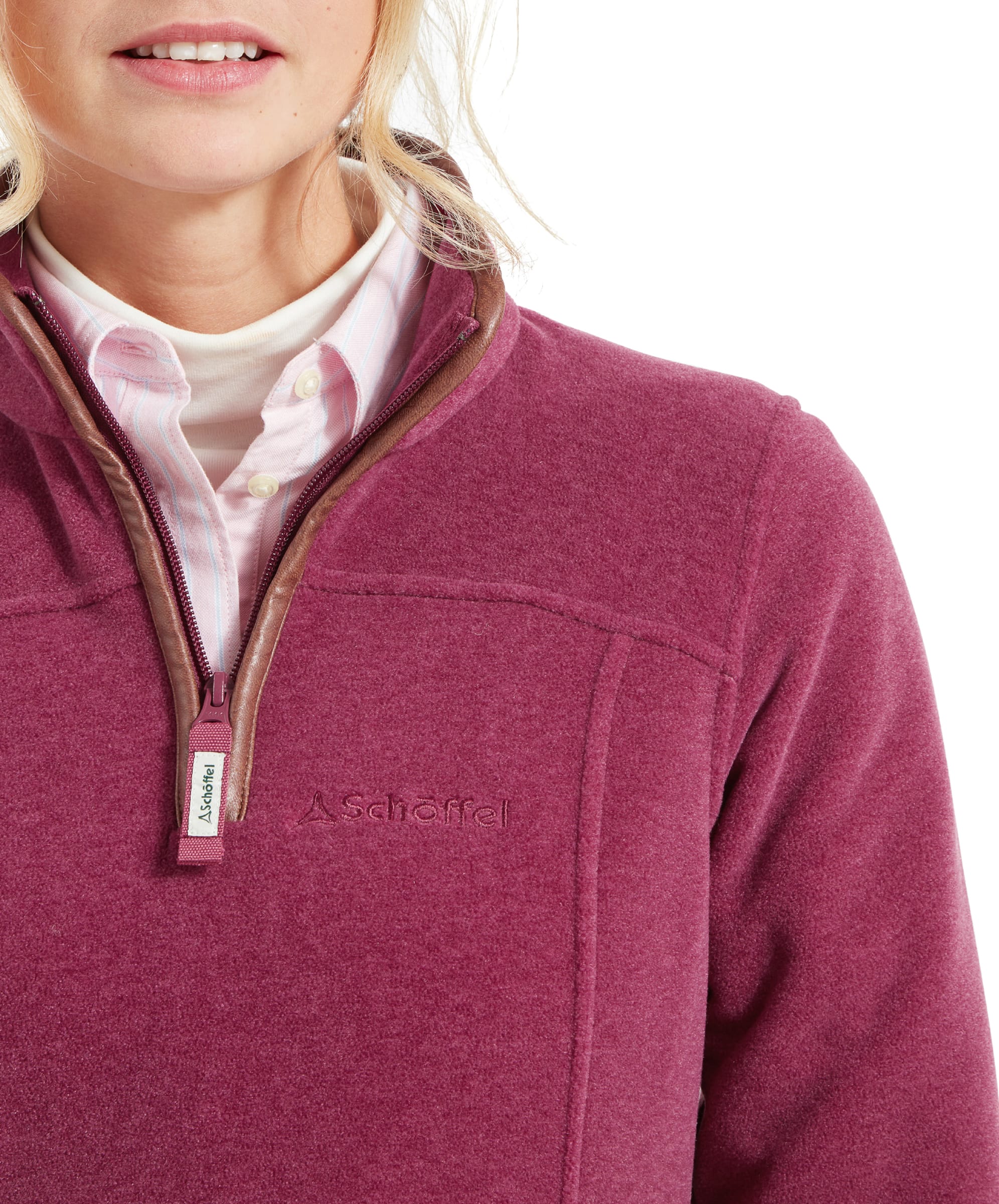 This image focuses on the front of the Schöffel Tilton 1/4 Zip Fleece for Women in Pink, showing the brown trim around the collar and zipper. The high collar is slightly open, revealing the light pink button-up shirt underneath. The brand’s logo is subtly embroidered on the chest, and the zipper pull is labeled with the brand name, highlighting the quality and attention to detail.