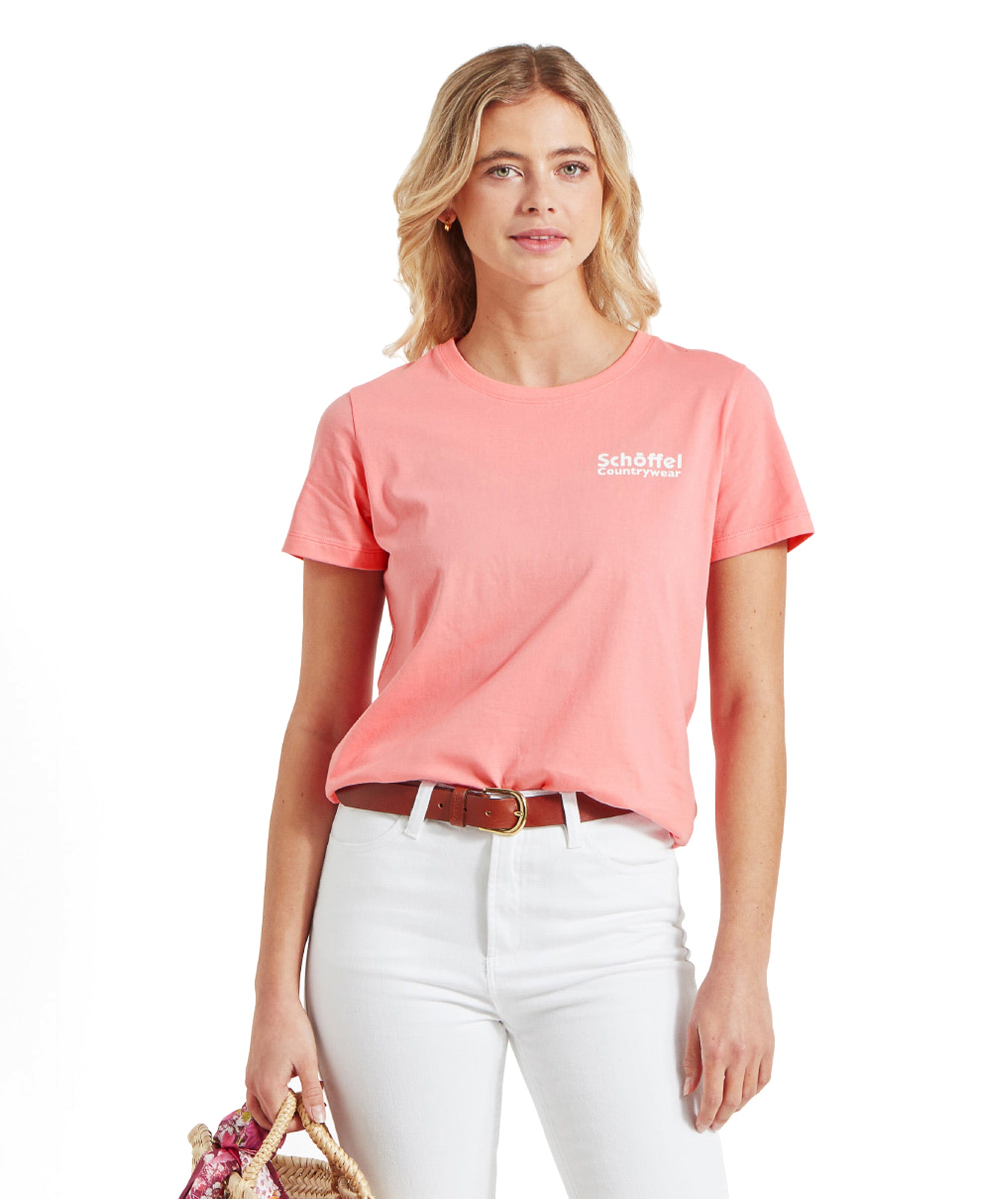 A model wearing a Schöffel Torre T Shirt for Women in Pink with white text on the chest, paired with white jeans and a brown belt, holding a straw bag.