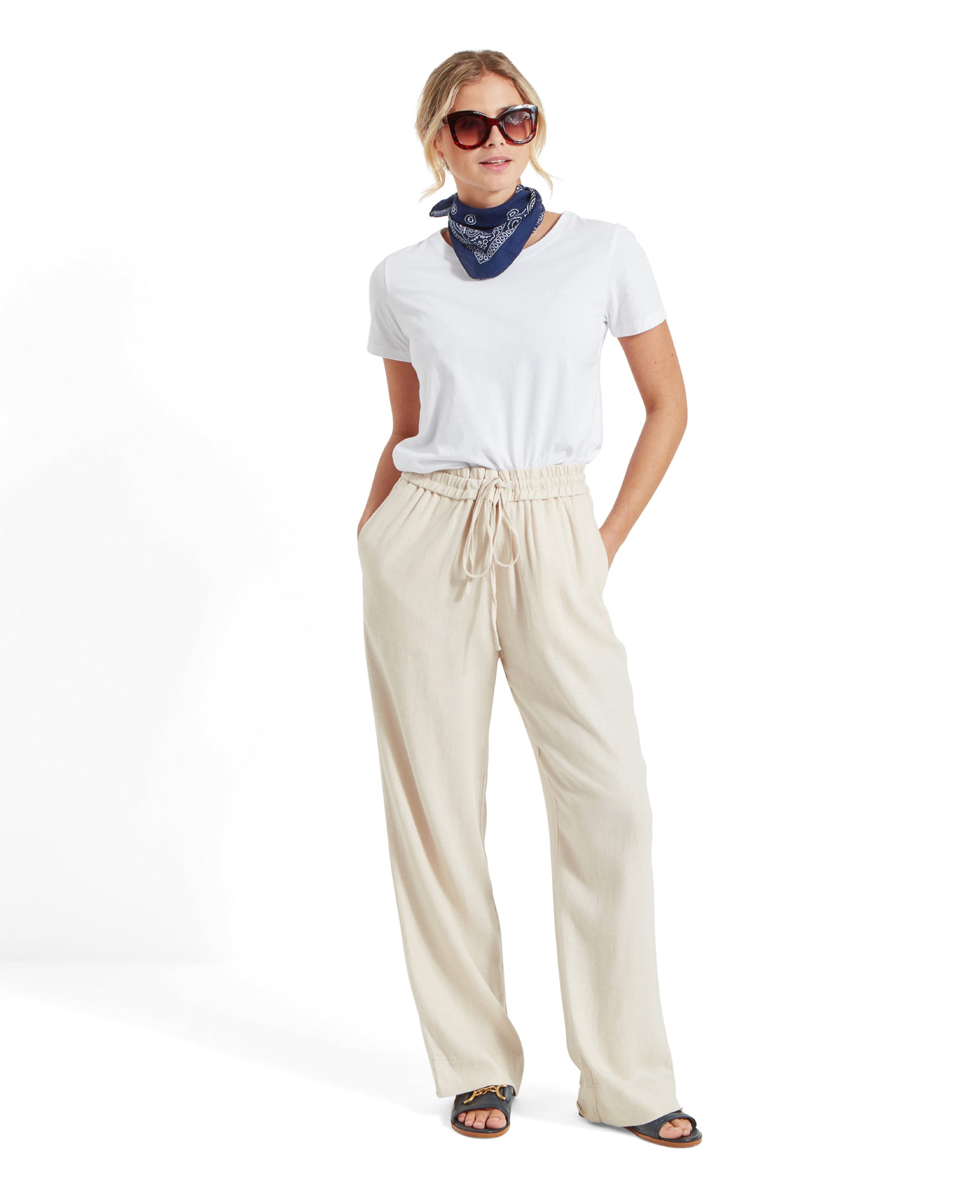 Full-body view of the woman in the Schöffel Tresco T Shirt for Women in White, beige wide-leg trousers, blue bandana, and sunglasses, standing and smiling.