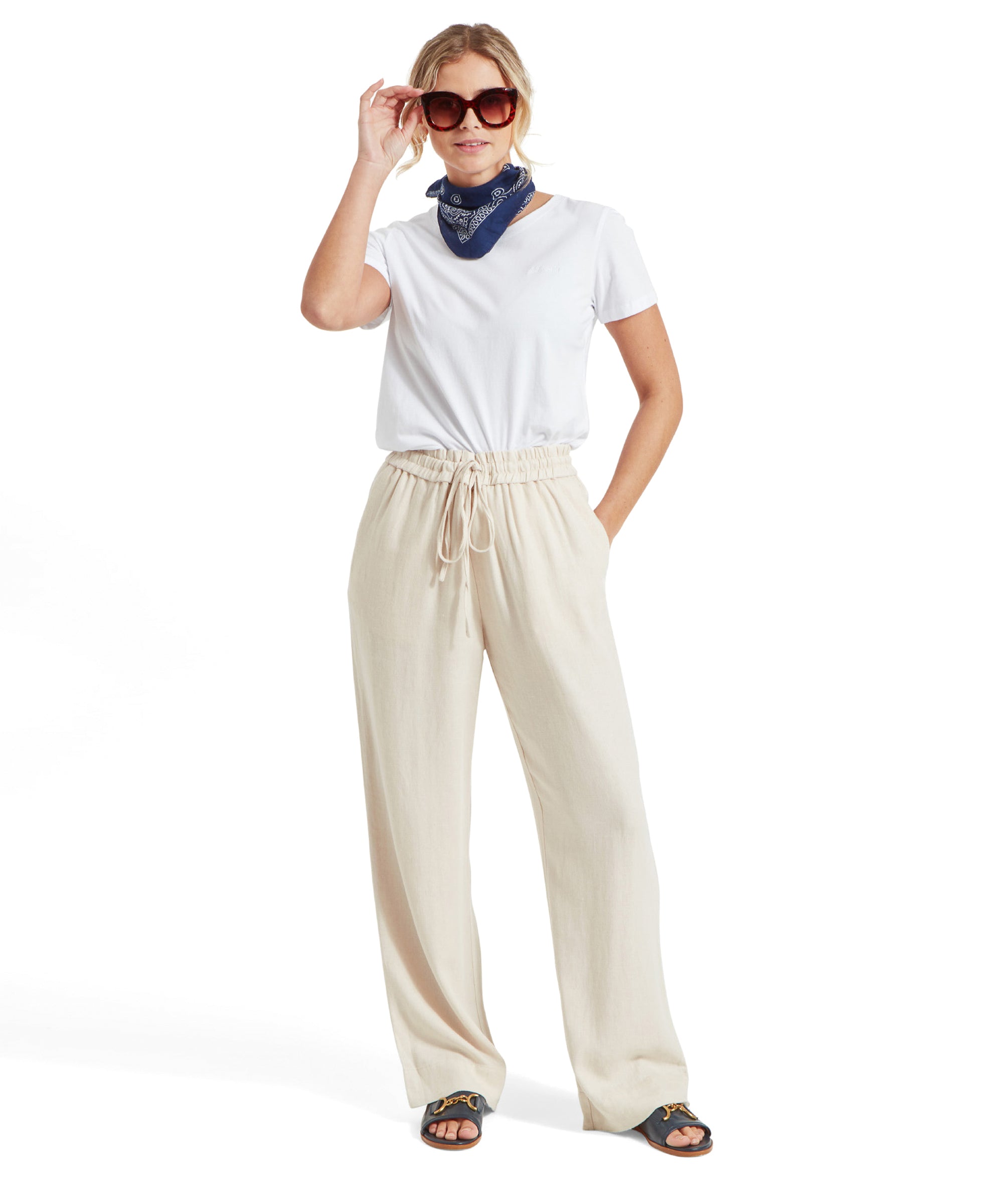 Full-body view of the woman in the white Tresco T-shirt, beige wide-leg trousers, blue bandana, and sunglasses, standing and holding her sunglasses.