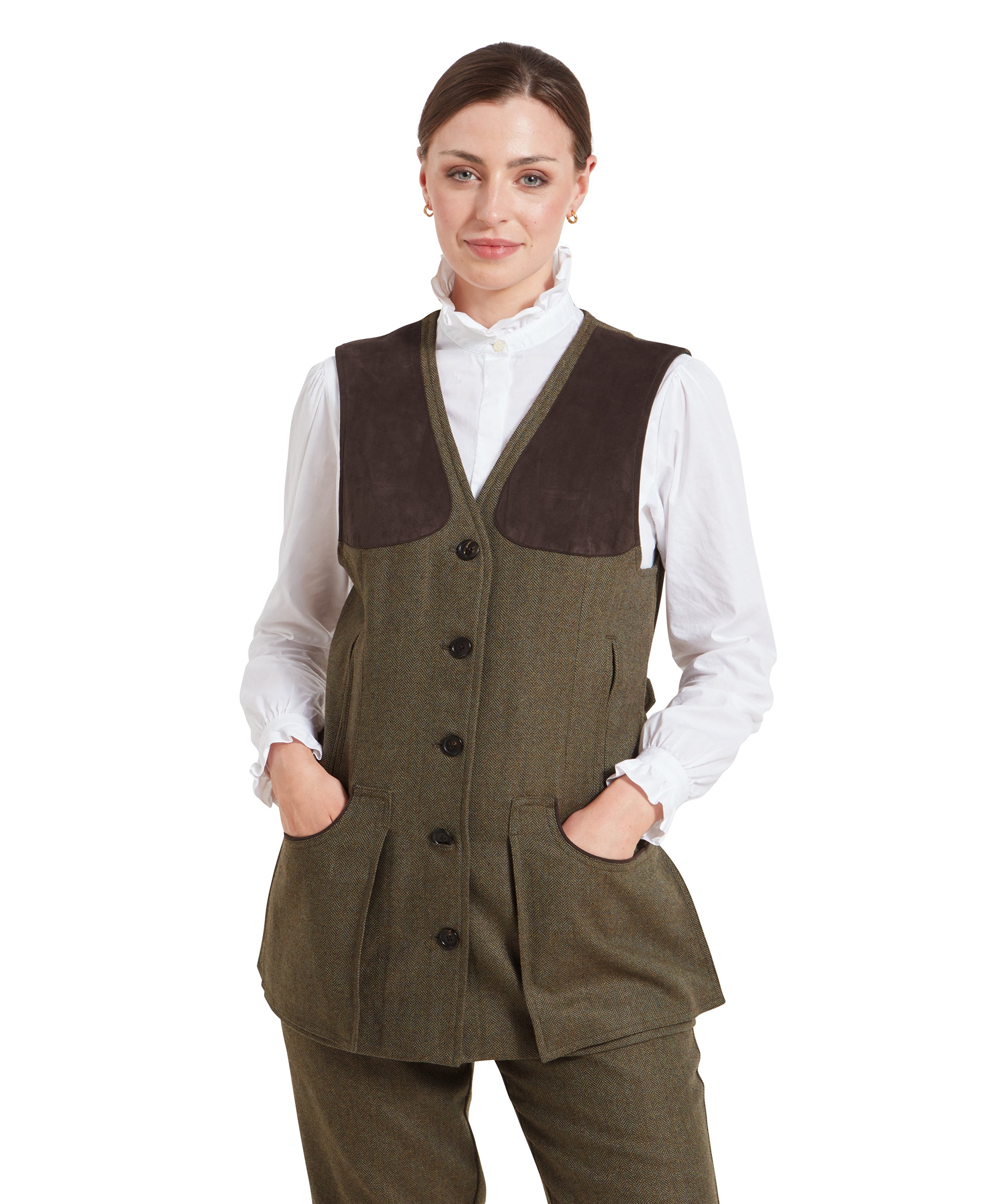 A woman wearing a Schöffel Women's Tweed Shooting Vest for Womens in Green with a brown shoulder patch, posing for a front-facing portrait. The vest is buttoned down with four large buttons and features two spacious front pockets. The model pairs the vest with a white blouse with ruffled cuffs and small gold hoop earrings. She has her hair neatly pulled back and is smiling slightly.