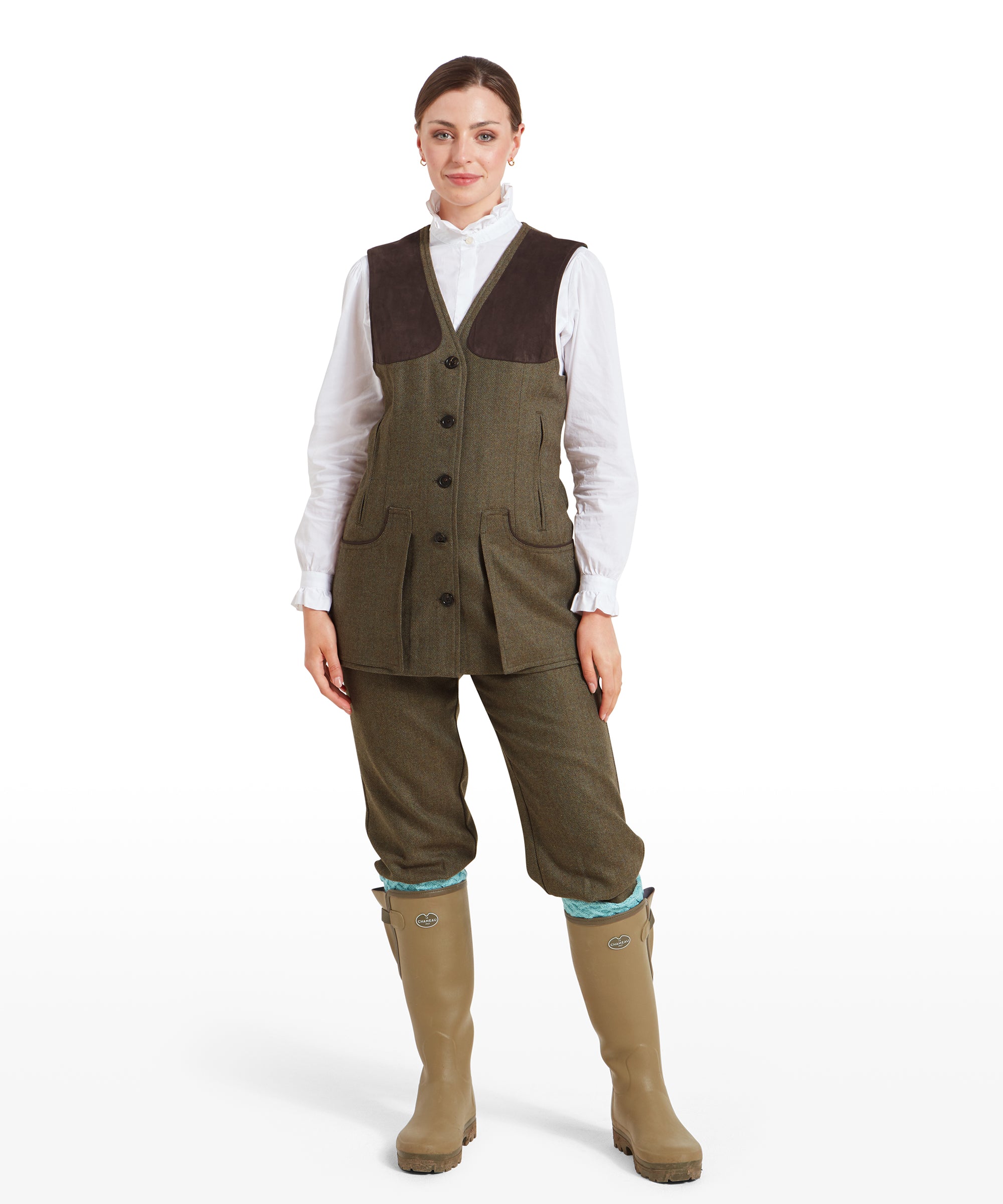 The same woman is shown in a full-body shot, wearing the tweed shooting vest over matching Schöffel Women's Tweed Shooting Vest for Womens in Green and beige Wellington boots. She stands facing forward with her hands resting casually in her pockets, highlighting the practical and traditional outdoor outfit.