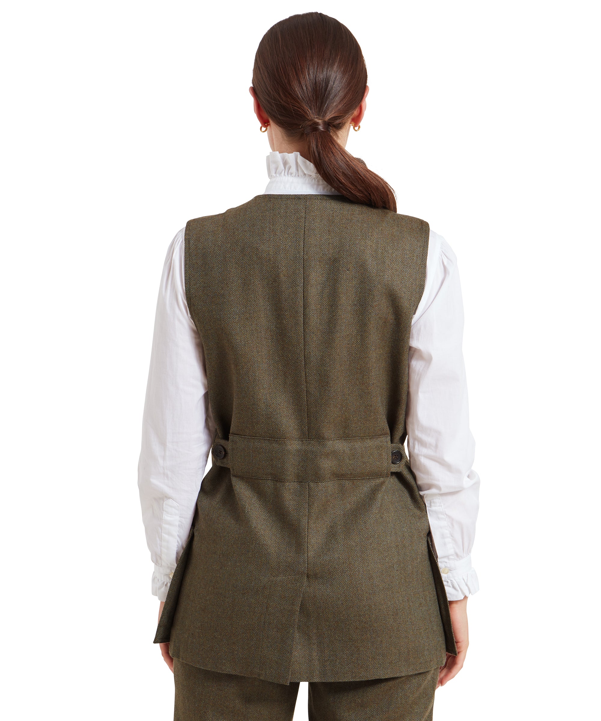 A rear view of the woman wearing the Schöffel Women's Tweed Shooting Vest for Womens in Green, showing the back details. The vest features a belt with adjustable buttons at the waist, providing a fitted appearance. Her hair is tied back into a low ponytail, complementing the clean, structured look of the outfit.