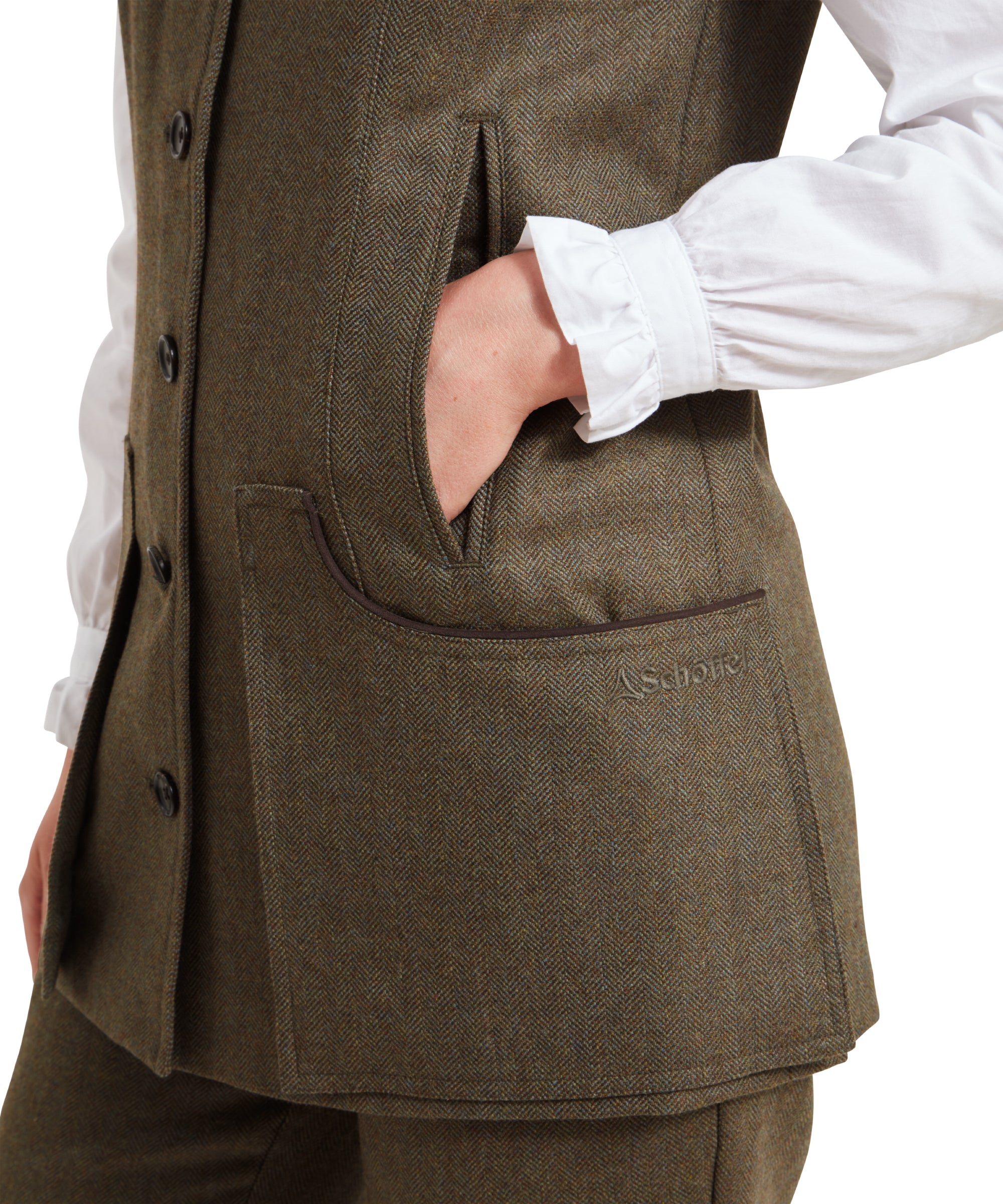 A close-up shot of one of the front pockets of the Schöffel Women's Tweed Shooting Vest for Womens in Green, with the woman’s hand casually placed inside. The pocket is lined with a brown trim, and the subtle herringbone pattern of the tweed fabric is clearly visible. The "Schöffel" logo is embroidered on the pocket, adding a refined touch to the vest's classic design.