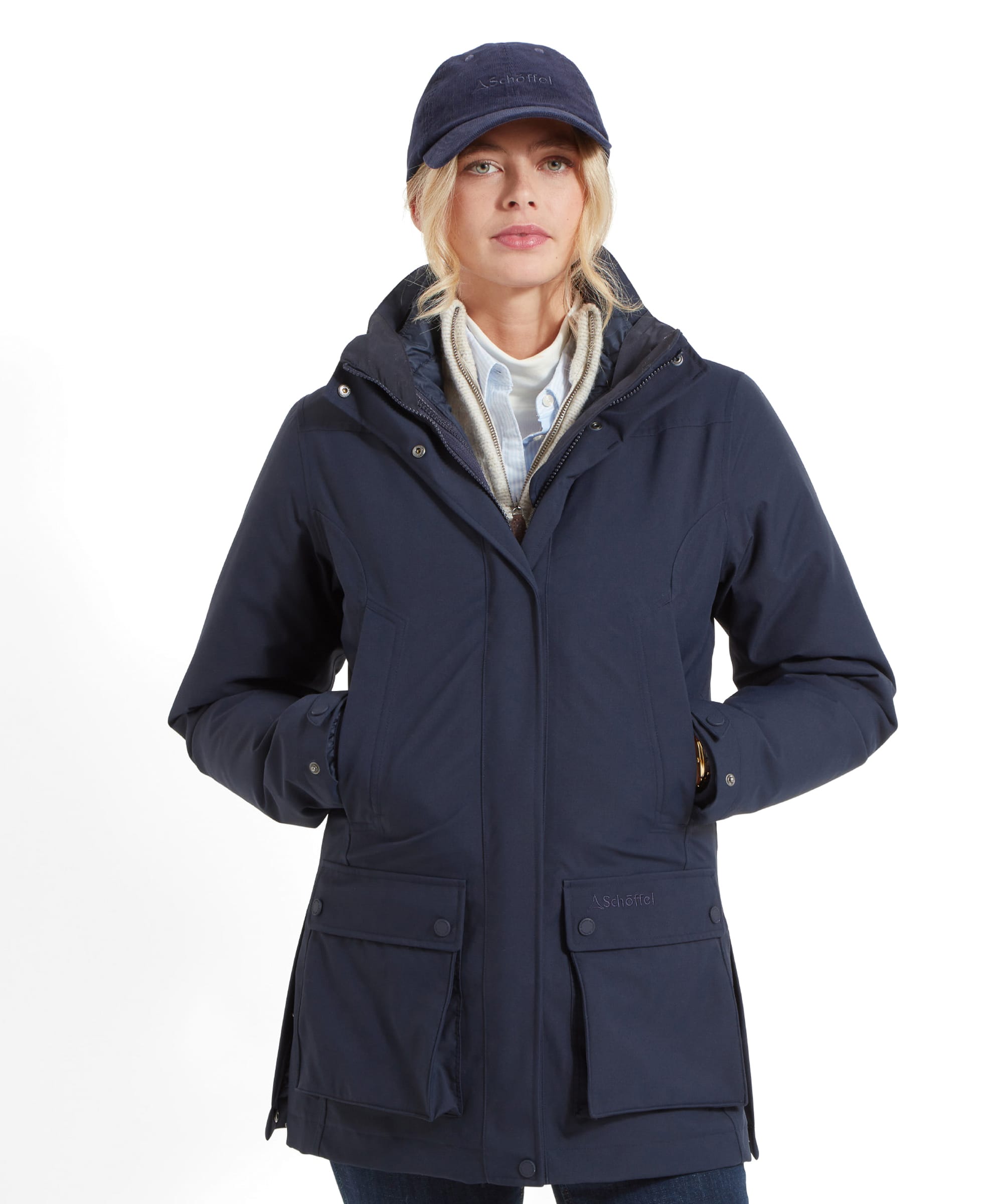 A woman wearing a Schöffel Uppingham 3-in-1 Coat for Women in Blue, standing with her hands in the coat pockets. She is also wearing a matching navy-blue baseball cap. The coat has a high collar and appears to be well-insulated, suitable for cold weather. The woman has blonde hair pulled back into a ponytail and is wearing a light-coloured shirt underneath the coat.