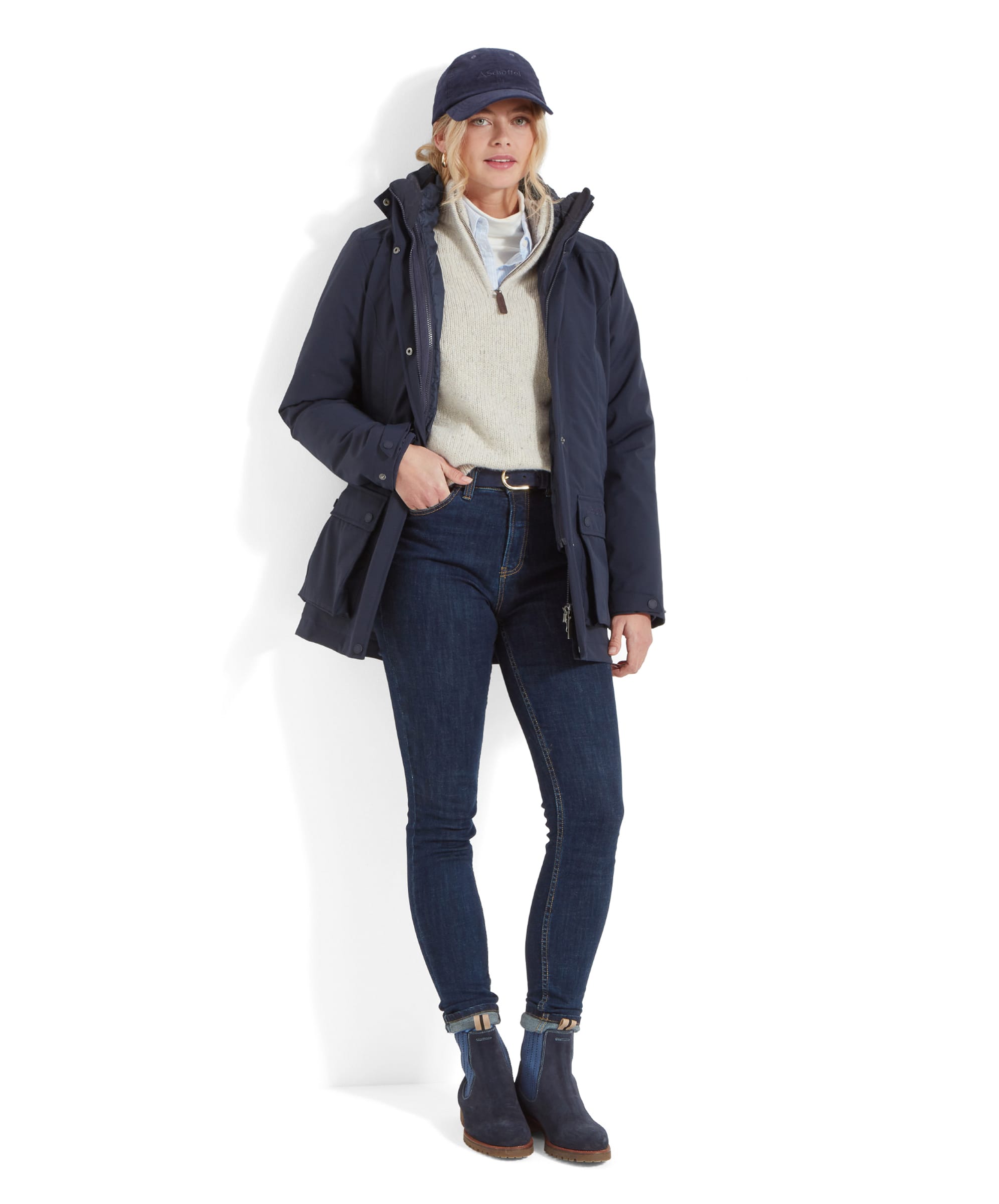 A woman is standing against a white background, dressed in a casual, layered winter outfit. She wears a Schöffel Uppingham 3-in-1 Coat for Women in Blue over a light beige sweater and a light blue collared shirt. Her slim-fit dark denim jeans are tucked into navy-blue ankle boots with light brown soles, and she completes the look with a matching navy-blue baseball cap. The woman has blonde hair pulled back in a low ponytail, and she poses with one hand in her coat pocket, exuding a relaxed and cosy style.