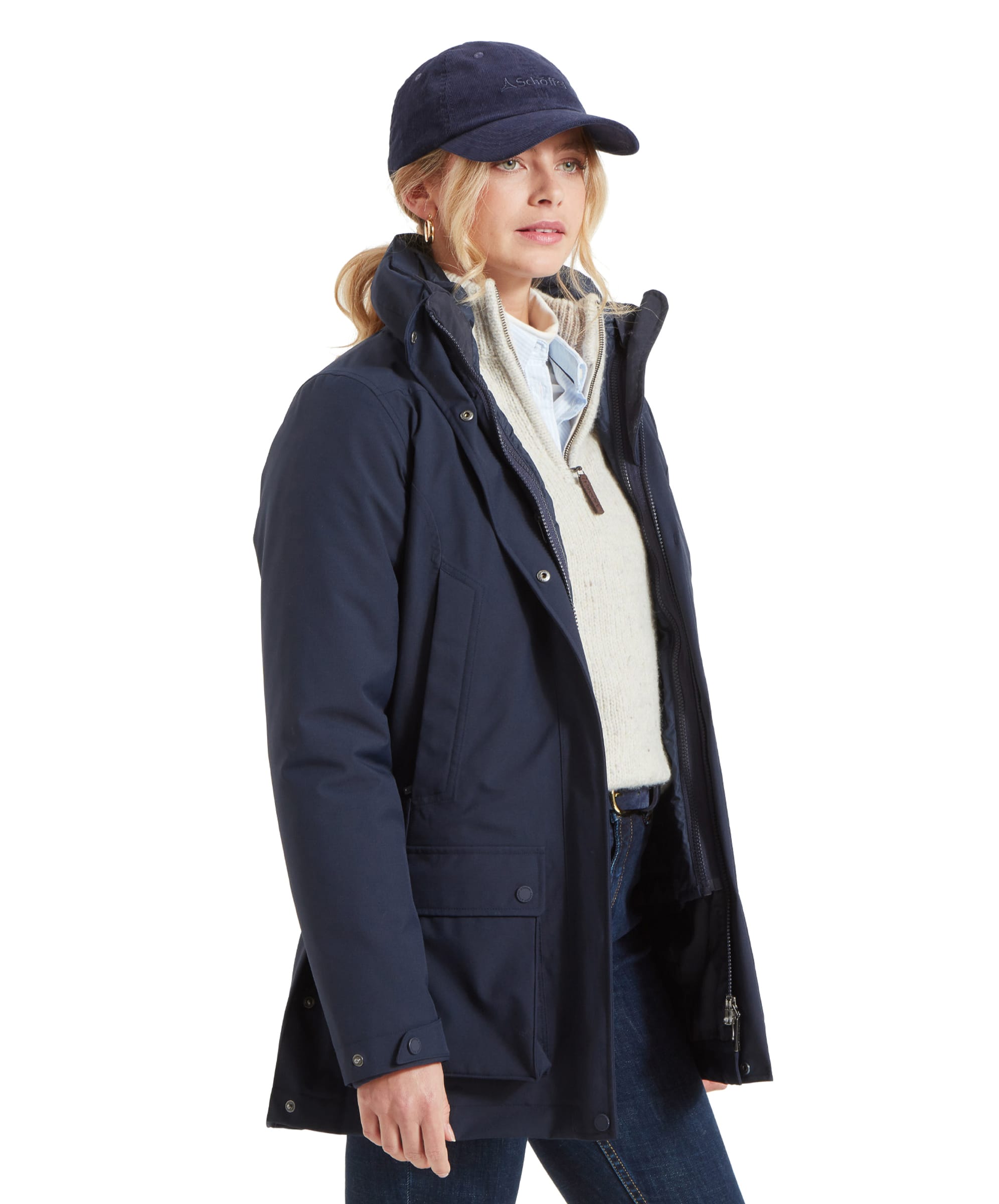 A side profile of the same woman in the Schöffel Uppingham 3-in-1 Coat for Women in Blue and matching baseball cap. She stands with her hands in the pockets, showing the coat’s clean, sleek lines and detailing. Her blonde hair is visible, styled in a ponytail, and she has a relaxed, casual posture.