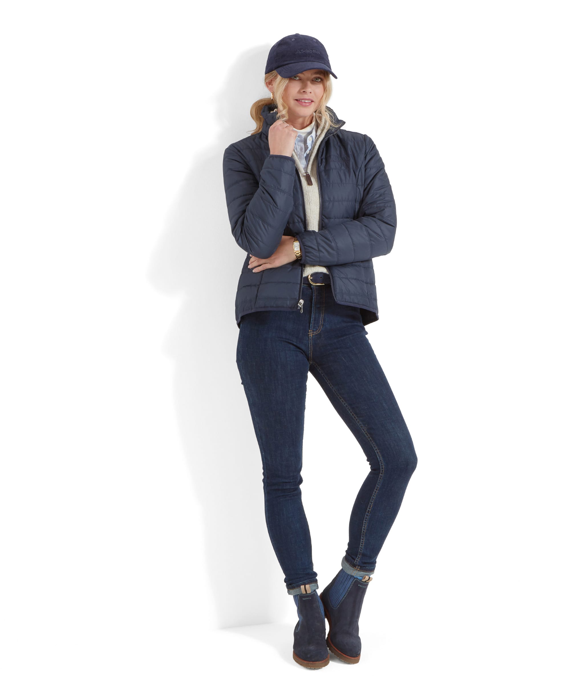 The woman is dressed in a casual outfit, wearing the lightweight quilted insert from the Schöffel Uppingham 3-in-1 Coat for Women in Blue, slim-fit jeans, and navy-blue ankle boots. She stands with one leg crossed over the other and her arms loosely folded, leaning slightly against a white background. She also wears the same navy-blue baseball cap, looking relaxed and stylish.