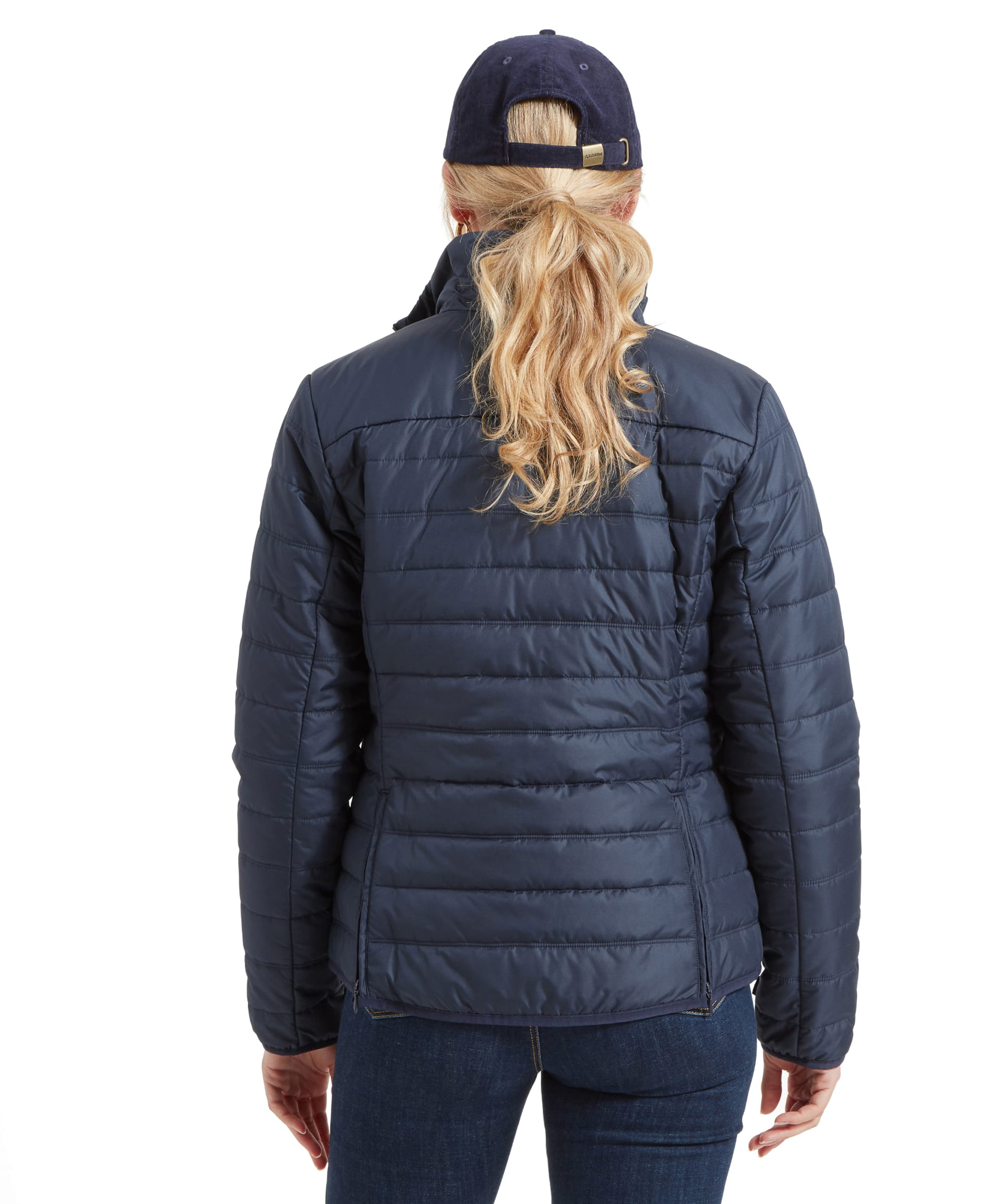 A rear view of the woman wearing the lightweight quilted insert from the Schöffel Uppingham 3-in-1 Coat for Women in Blue. Her blonde hair is tied back in a low ponytail, and she wears the same navy-blue baseball cap. The jacket’s horizontal quilting is visible, emphasizing the jacket’s warmth and structure.