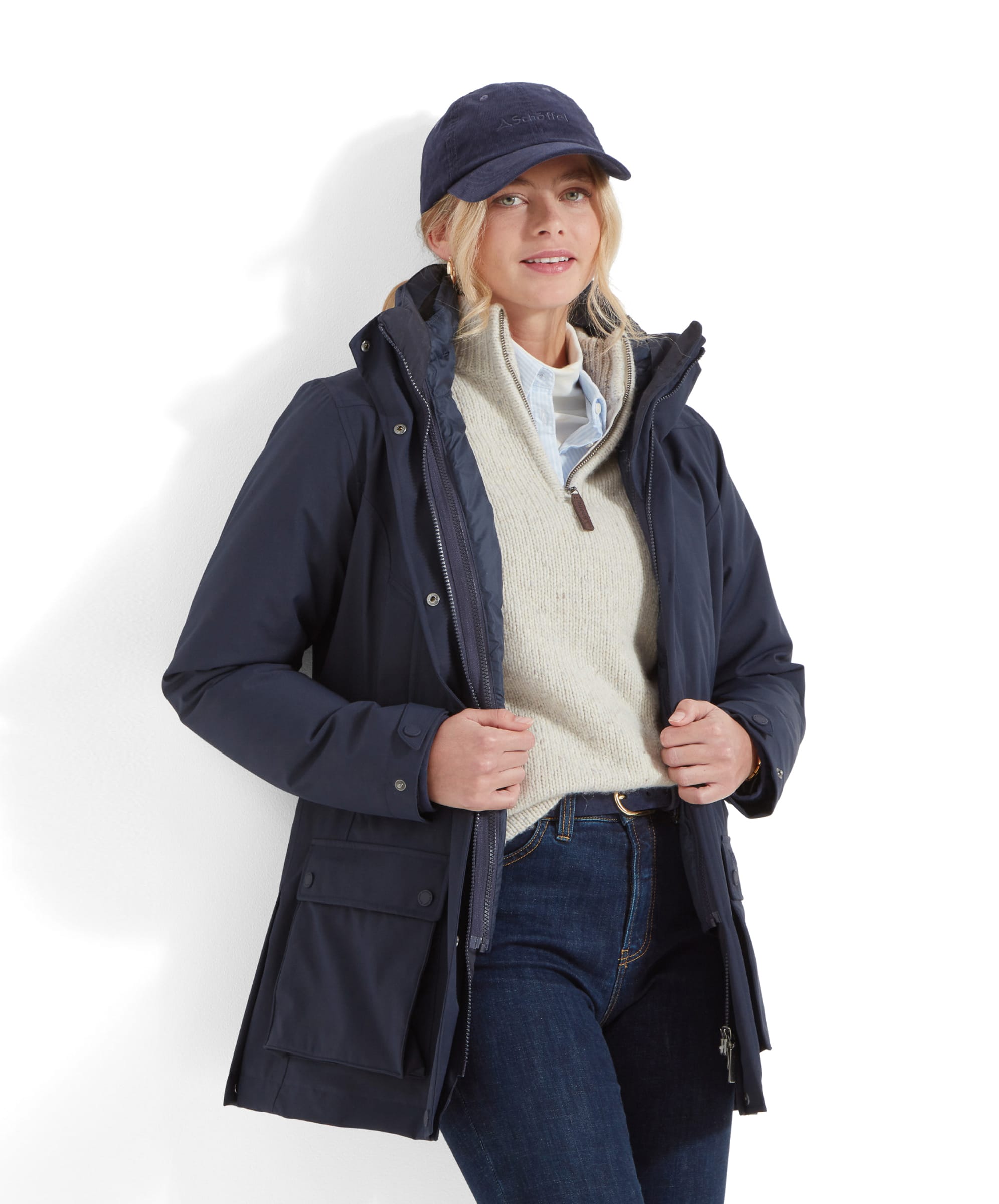 The woman standing slightly angled, wearing the Schöffel Uppingham 3-in-1 Coat for Women in Blue, with the front partially unzipped to show a beige sweater underneath. She has her hands in her coat pockets and looks relaxed. She also wears slim-fit jeans and a navy-blue baseball cap.