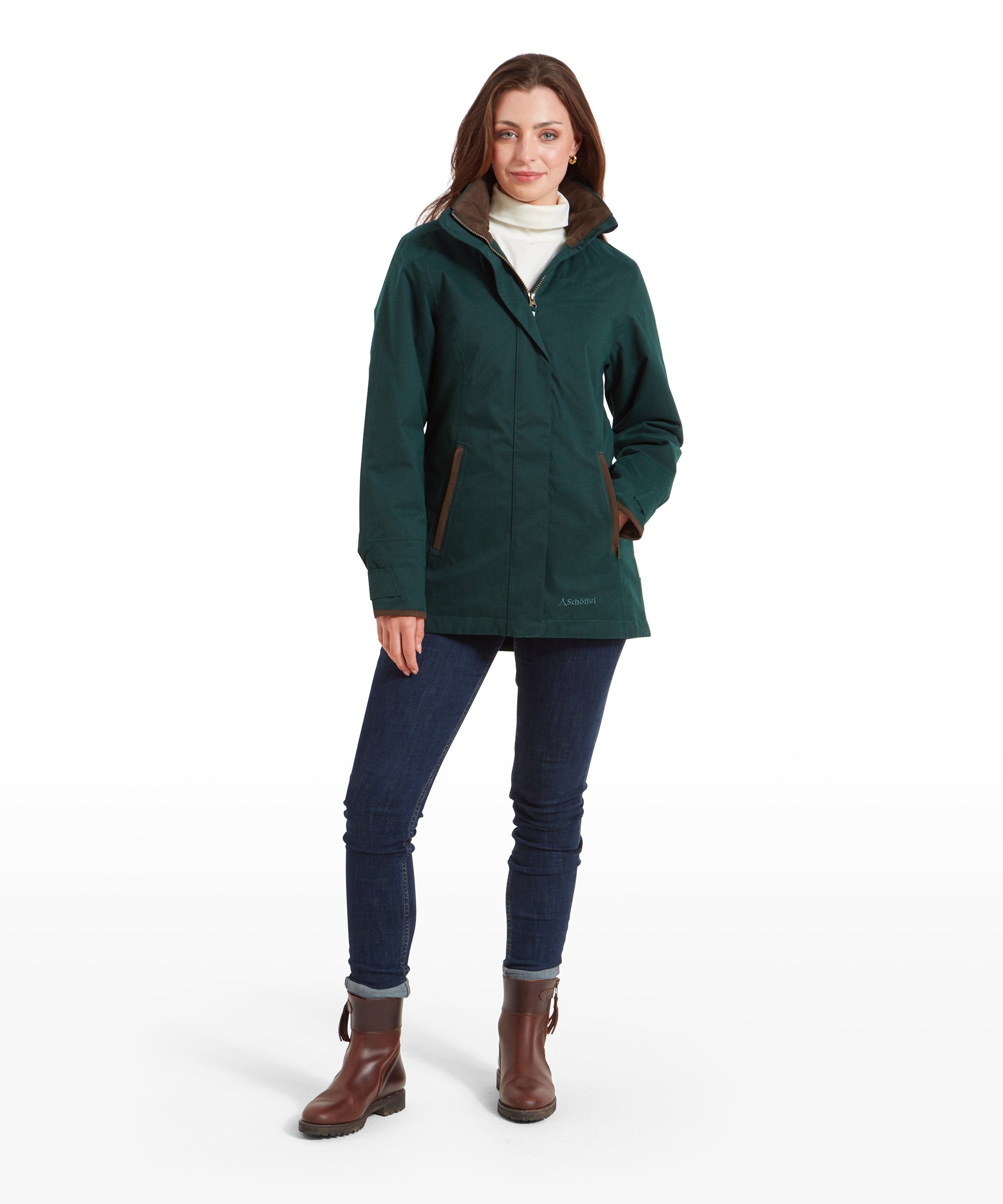 The same woman stands confidently looking at the camera, wearing the Schöffel Wakerley Jacket for Women in Green full zipper. She partners the coat with a cream turtleneck top, dark denim jeans, slightly rolled up at the hem and accessorised with brown boots.