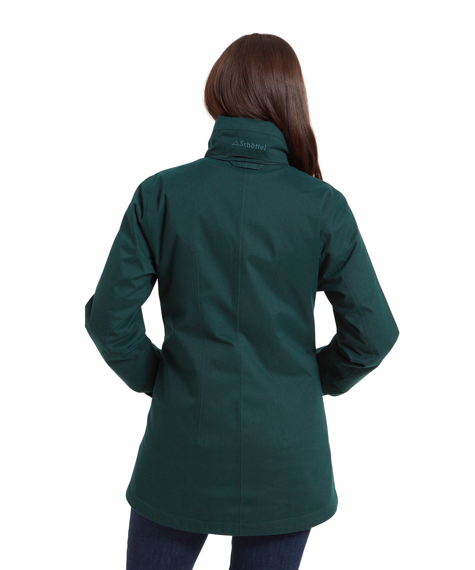 Rear view of the woman wearing the Schöffel Wakerley Jacket for Women in Green, this time with her hands in both the front pockets and her brunette hair tucked into the collar area. On the back of the collar a Schöffel embroidered logo can be seen in the hood pocket and the fit of the jacket can be clearly seen from this angle.