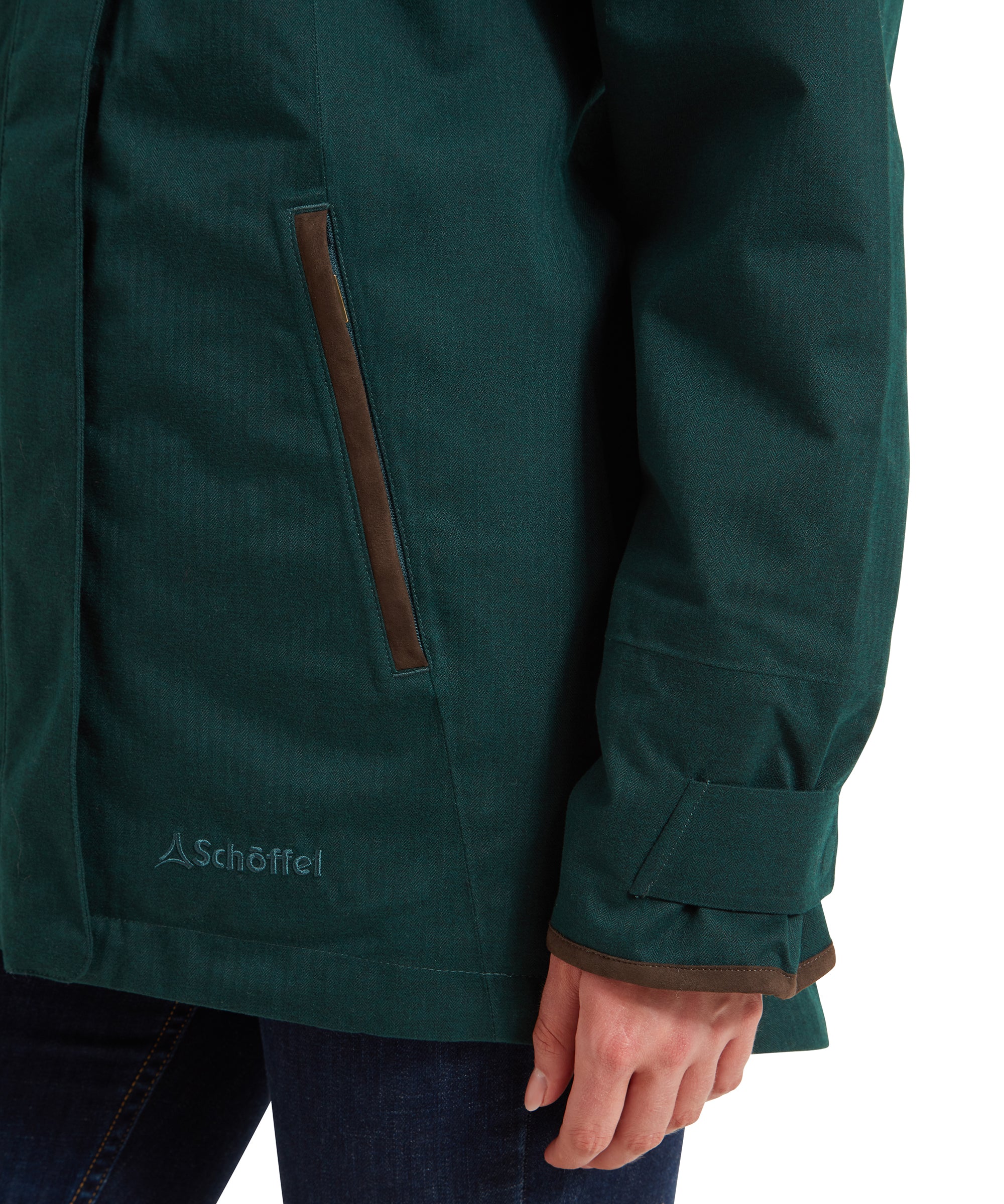 A close-up of the Schöffel Wakerley Jacket for Women in Green hem and adjustable cuff. You can see a Schöffel embroidered patch to the side of the lower zip. The edges of the coat cuff and pocket has been edged with the brown suedette fabric.