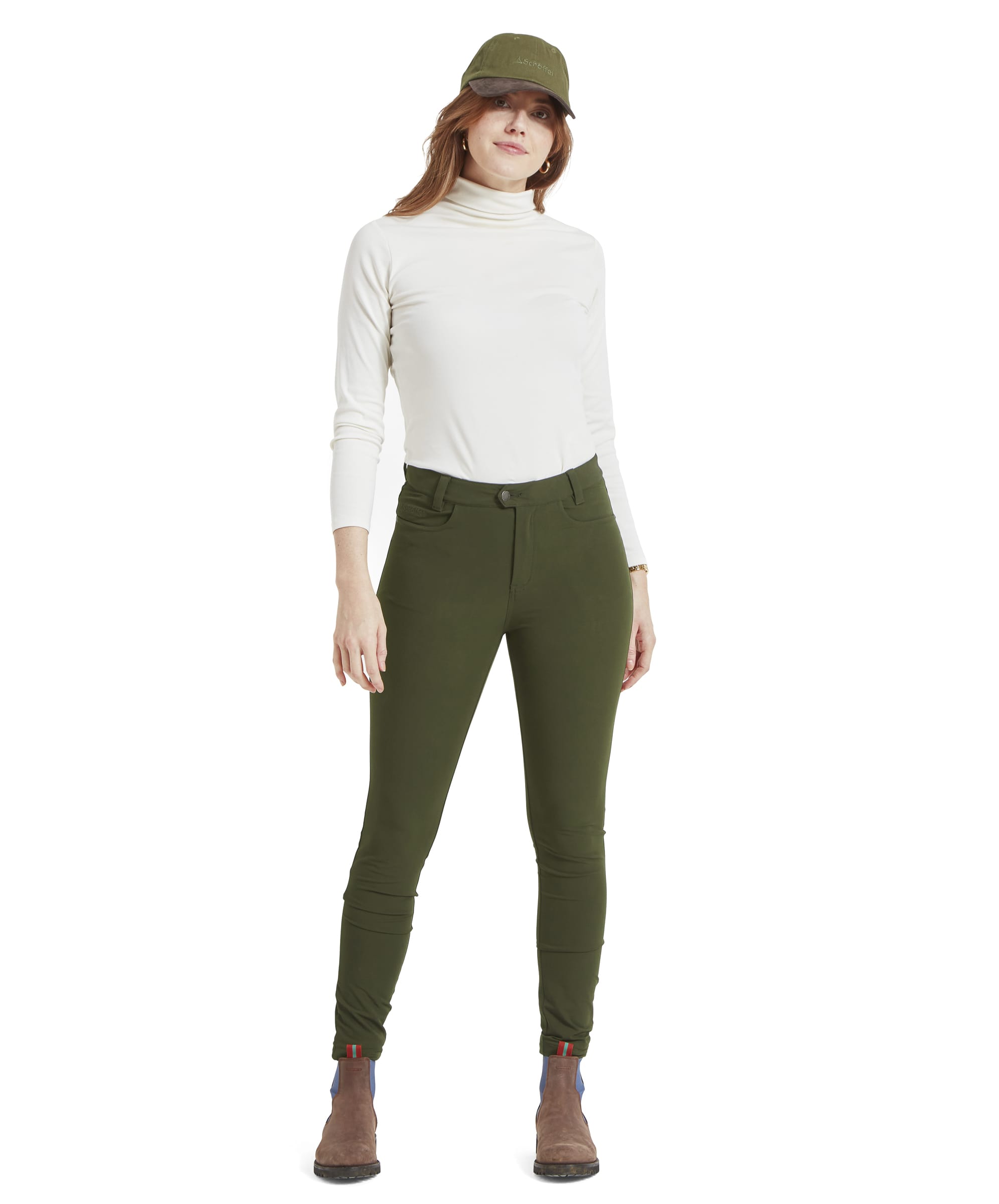 The model is standing in a relaxed pose, wearing a white long-sleeved top tucked into Schöffel Whitwell Water Repellent Trouser for Women in Green. She is also wearing brown boots with blue elastic side panels and a green cap. The image shows the entire outfit from head to toe, emphasizing the fit and cut of the trousers.