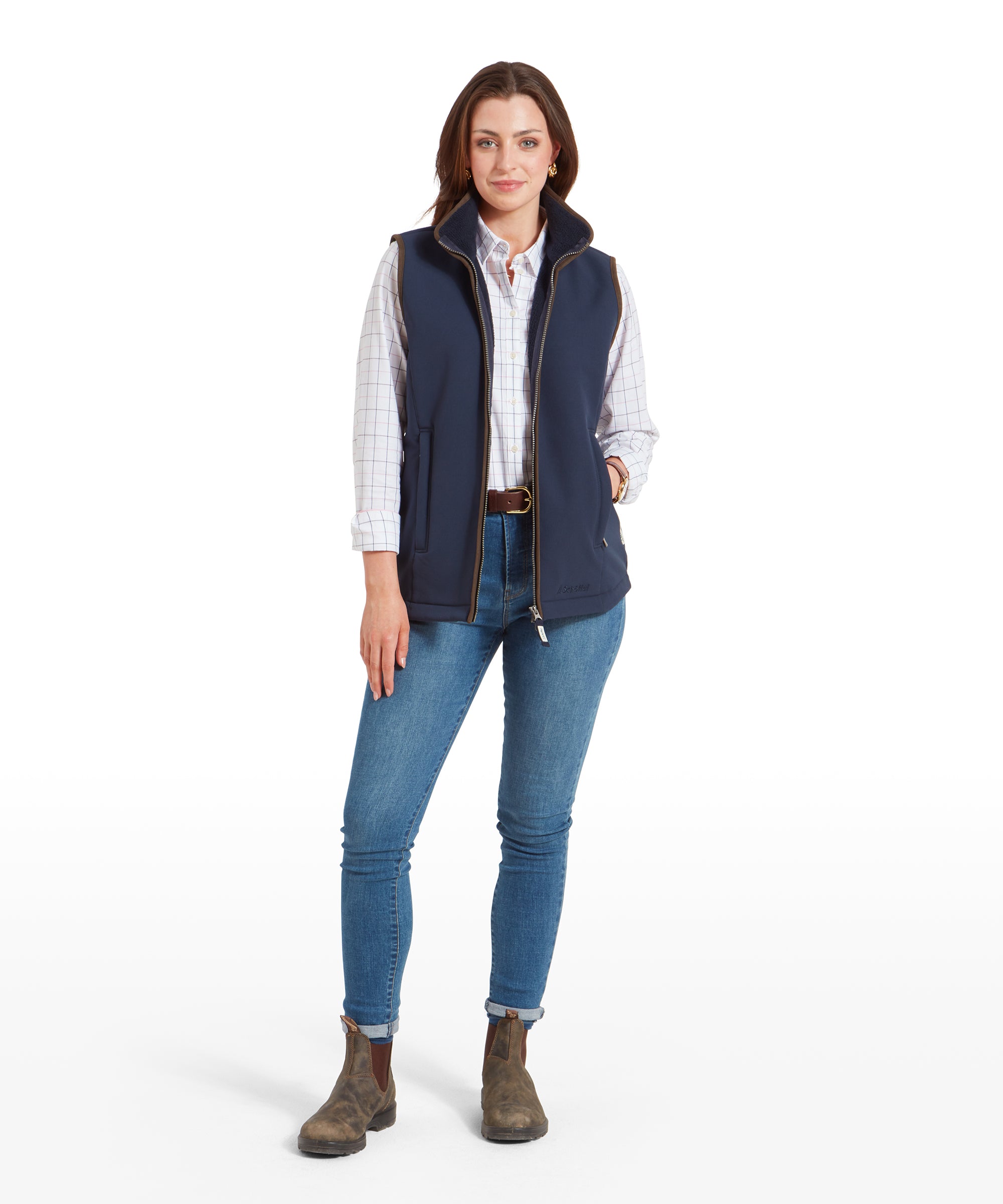 A full-body shot of the woman wearing the Schöffel Belton Gilet for Women in Navy, paired with blue jeans and brown boots. She is standing with her hands in her pockets and looking directly at the camera.