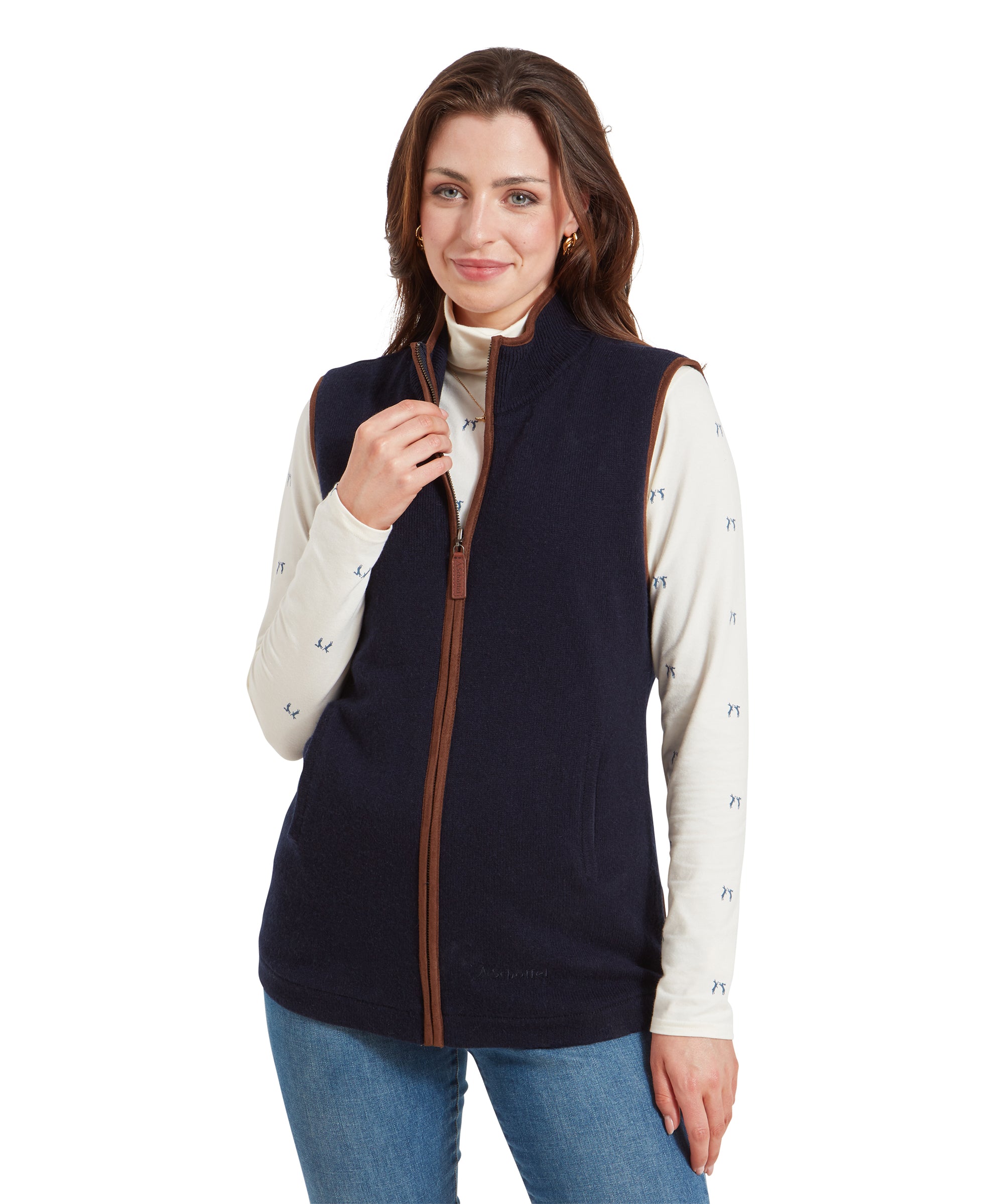 A woman wearing a Schöffel Women's Lambswool Aerobloc Gilet for Womens in Navy over a cream long-sleeve top with a subtle print, paired with light blue jeans. She is smiling and standing with her hand near the zipper of the gilet.