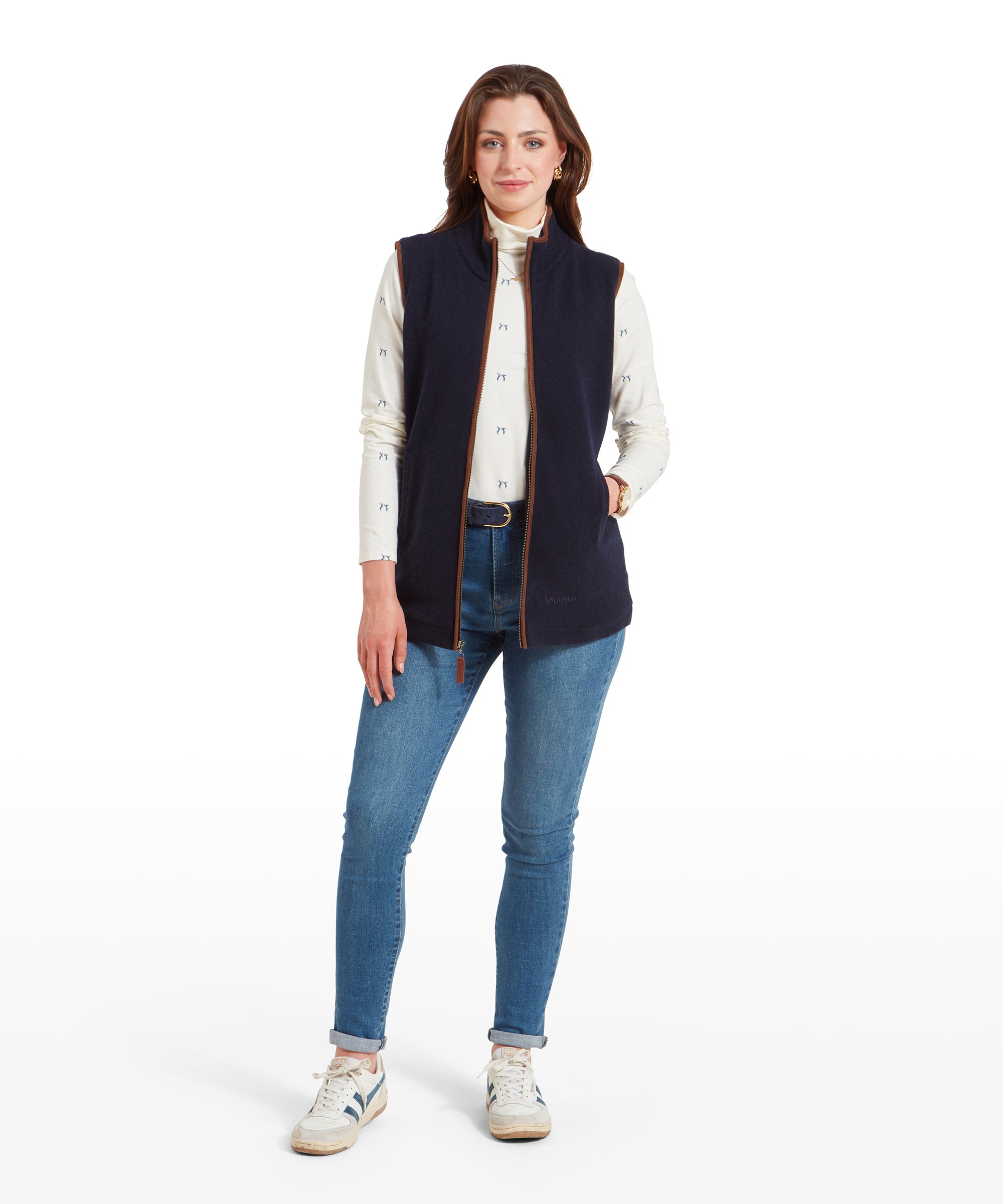 A full-body shot of a woman wearing a Schöffel Women's Lambswool Aerobloc Gilet for Womens in Navy over a cream long-sleeve top with small printed designs and light blue jeans. She is standing casually with her hands in her pockets, wearing white trainers.