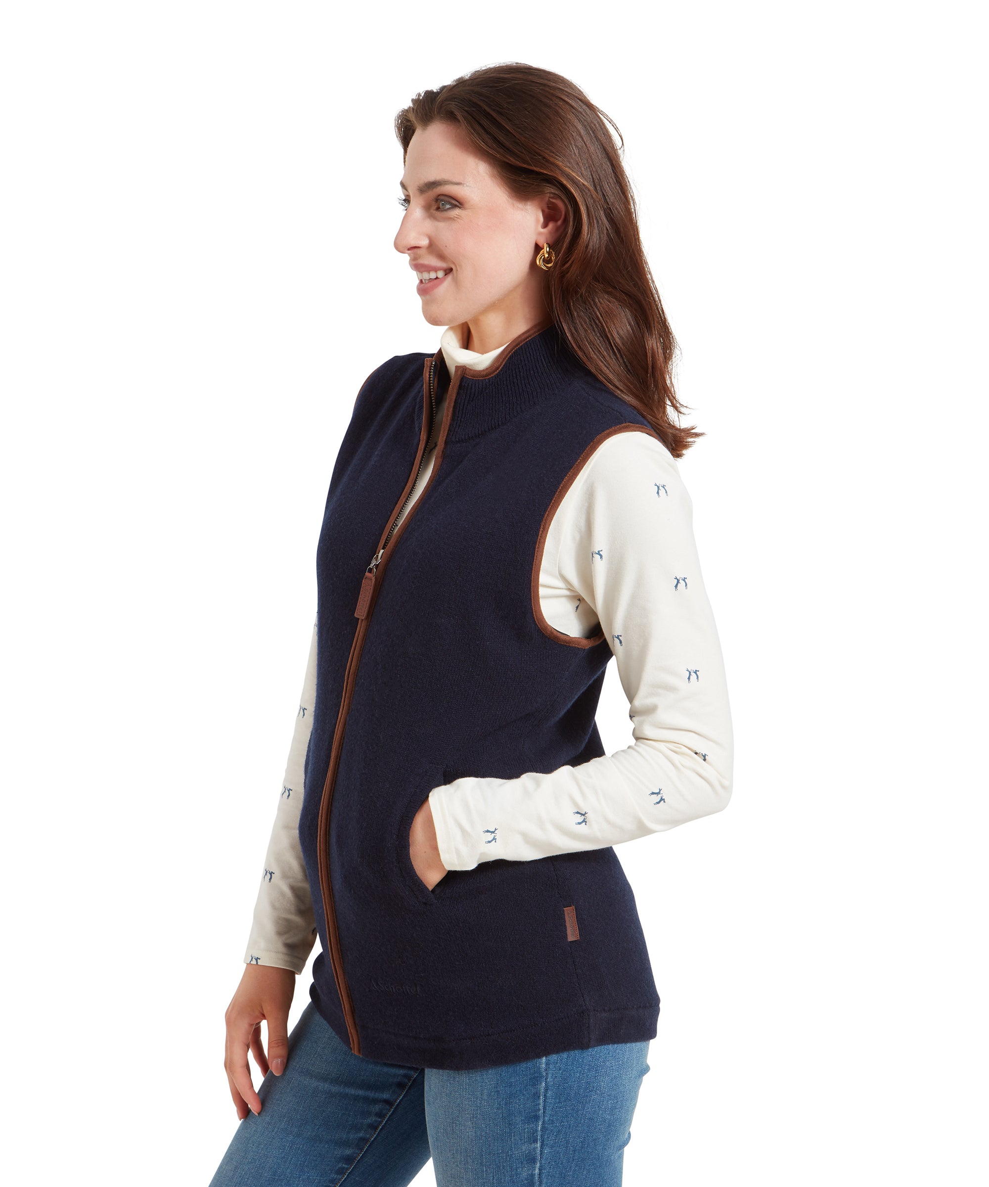 A side profile of a woman wearing a Schöffel Women's Lambswool Aerobloc Gilet for Womens in Navy with brown trim over a cream top with small blue prints, paired with light blue jeans. Her hands are in her pockets as she smiles.