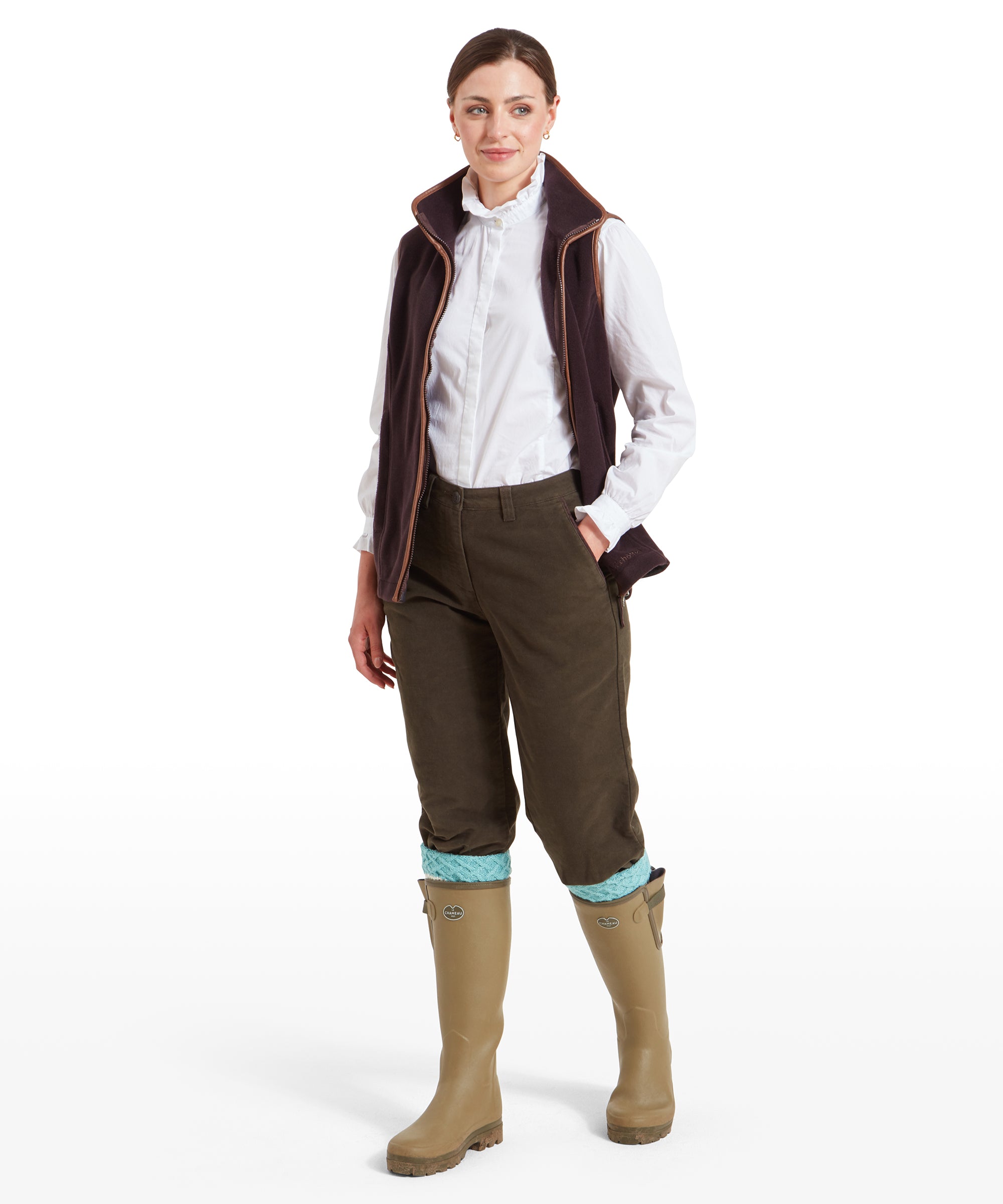 A woman dressed in Schöffel Moleskin Breeks for Womens in Green paired with a white shirt and brown fleece gilet. The breeks are tucked into beige wellington boots with light blue knit socks. She stands in a relaxed pose with her hands in her pockets.